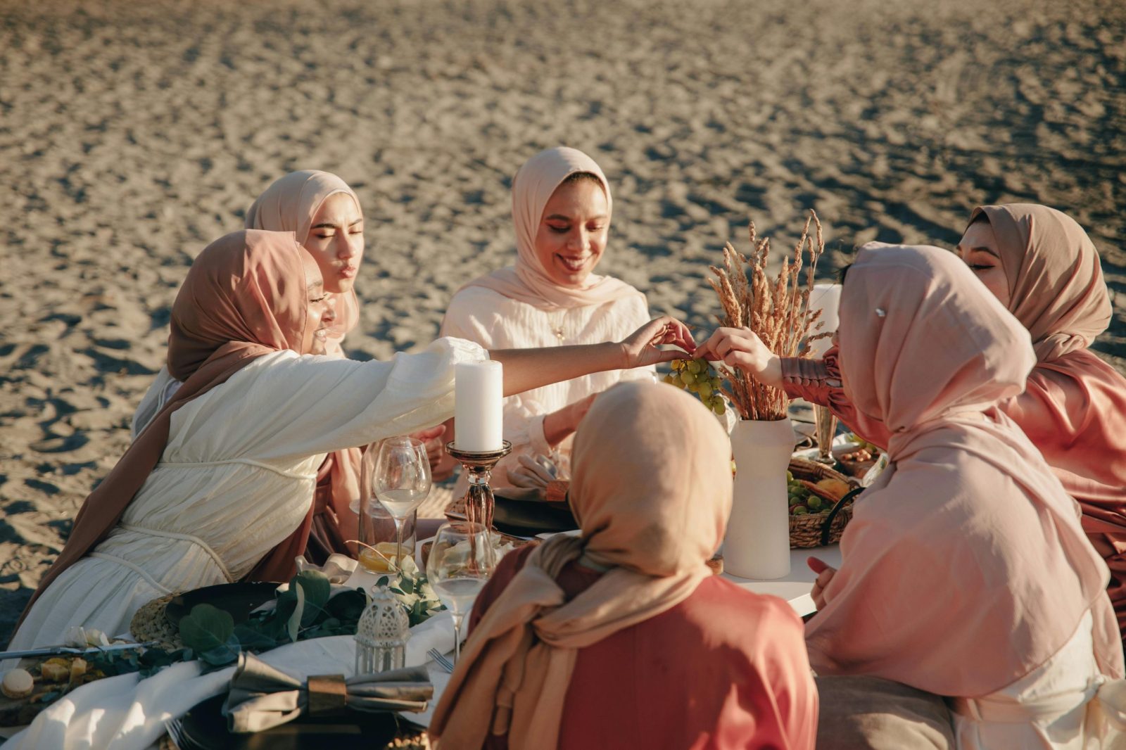 A group of women in hijabs enjoying a beach picnic with food and drinks on a sunny day.